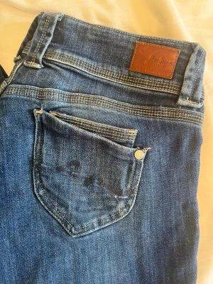 Blå straight fit jeans från Pepe Jeans - Snygga blå straight fit jeans från Pepe Jeans med klassisk femficksdesign och dubbla knappar i midjan. Jeansen har en mörk tvätt och detaljerade sömmar, samt läderpatch med logga bak. Perfekta för en trendig och avslappnad look.