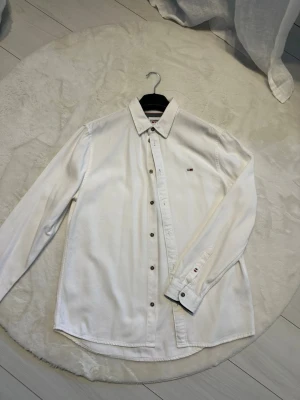 Vit skjorta från Tommy Jeans - Säljer en jättefin vit långärmad skjorta från Tommy Jeans. Den är tillverkad i ett lite tjockare, mjukt och jätteskönt bomullstyg med snygga bruna knappar och den klassiska loggan på bröstet. Skjortan är i bra skick då den bara är använd ett fåtal gånger. Skickas snabbt, hör av dig om du undrar över något eller vill ha mått! 