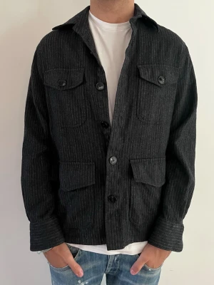 Svart overshirt med smala ränder - Stilren svart overshirt med tunna vita kritstrecksränder, fyra fickor med lock och knappar framtill. Rak passform och klassisk krage. Perfekt att bära över en t-shirt för en avslappnad men snygg look. Materialet ser ut att vara mjukt och lite tjockare, troligen bomull eller ullmix.