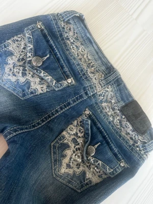 Blå bootcut jeans med spets och paljetter - Snygga blå bootcut jeans med dekorativa bakfickor i spets och paljettdetaljer. Jeansen har slitningar och unika knappar på fickorna. 