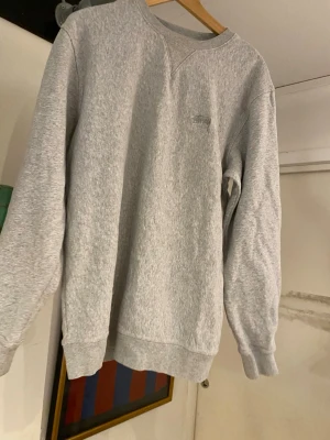 Grå sweatshirt från Stüssy - Klassisk grå sweatshirt från Stüssy med broderad logga på bröstet. Tröjan har rund halsringning, långa ärmar och ribbade muddar vid ärmslut och nederkant. Perfekt för en avslappnad streetwear-look.