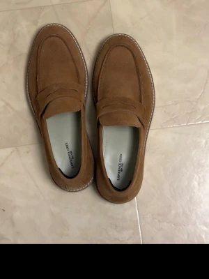 Bruna mockaloafers från Lawrence Grey - Snygga loafers i brun mocka från Lawrence Grey. Skorna har klassisk pennyloafers-design med dekorativ rem över vristen och vita kontrastsömmar runt sulan. Perfekta för en stilren och avslappnad look.