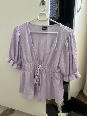Ljuslila blus  Gina Tricot - Söt ljuslila blus från Gina Tricot med djup v-ringning och knytband i midjan. Blusen har puffiga trekvartsärmar med volang vid ärmslut och är tillverkad i ett strukturerat, lätt tyg som ger en luftig känsla.