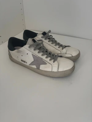 Golden goose vita sneakers med grå stjärna - Säljer ett par vita sneakers från Golden Goose med grå mockastjärna på sidan, grå skosnören och svart häl. Skorna har GGDB-logga och slitna detaljer för en cool, avslappnad look. Tillverkade i läder och mocka. 