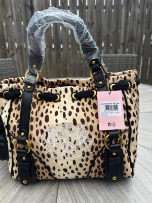 Juicy Couture leopardmönstrad handväska - Cool handväska från Juicy Couture med leopardmönster i beige och brunt. Väskan har svarta detaljer i läderimitation, guldfärgade metalldelar och ett broderat emblem framtill. Praktiska handtag och snygga spännen ger en trendig look.