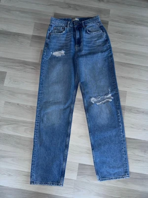 Blå jeans med slitningar - Snygga blå mom jeans från Gina Tricot med hög midja och raka ben. Jeansen har slitna detaljer på låret och knät för en trendig look. Klassisk femficksmodell i 100% bomull. Perfekta för dig som gillar en avslappnad och cool stil.