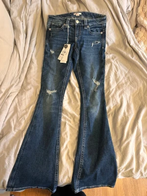 Blå bootcut jeans från Gina Tricot - Bootcut jeans i blå denim från Gina Tricot med slitningar på låren och knäna. Jeansen har klassisk femficksdesign, vita sömmar och är tillverkade i bomull med stretch. Perfekt för dig som gillar en trendig och avslappnad look.