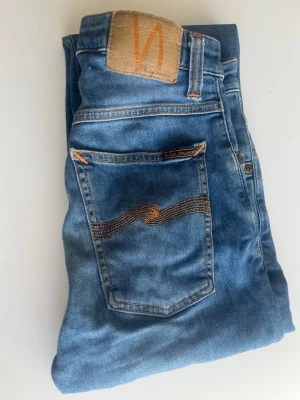 Nudie Jeans - Säljer ett par klassiska blå jeans från Nudie Jeans med snygga kontrastsömmar och ikonisk vågdetalj på bakfickorna. Jeansen har normal passform och är tillverkade i mjukt denimtyg. Det står 28/34 men de passar typ 31/32 skulle jag säga. Skicket är näst intill perfekt, inga skavanker finns. Skriv gärna vid frågor🙌