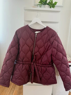 Vinröd quiltad pufferjacka H&M - Snygg vinröd pufferjacka från H&M med quiltat mönster och rund halsringning. Jackan har dragkedja framtill och ett matchande knytband i midjan. Använd en gång. Nypris: 549kr 