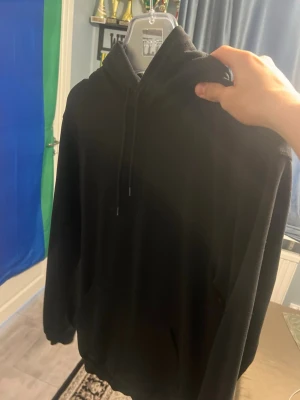Svart hoodie med huva och dragsko - En klassisk svart hoodie med huva och dragsko. Hoodien har en avslappnad passform, långa ärmar och en stor magficka framtill. Perfekt för chill dagar eller när du vill ha en enkel och stilren look. Materialet känns mjukt och bekvämt mot huden.