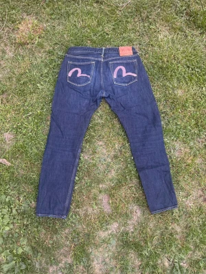 Evisu Jeans - Säljer ett par mörkblå jeans från Evisu Jeans med klassisk rak passform. Jeansen har Evisus ikoniska broderi på bakfickorna och en läderpatch i midjan. Tillverkade i slitstark denim av bomull. Perfekta för dig som gillar stilrena och tidlösa jeans.