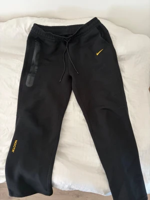 Svarta Nike mjukisbyxor med gul logga - Snygga svarta mjukisbyxor från Nike med gul swoosh och NOCTA-detalj på benet. Byxorna har dragsko i midjan, resår och en cool ficka med dragkedja på sidan. Perfekta för chill eller streetwear-stil.