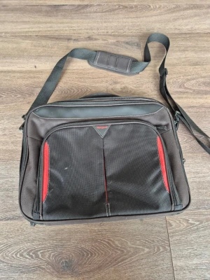 Svart datorväska med röd detalj - Snygg svart datorväska med röda detaljer och justerbar axelrem. Väskan har flera fack, vadderat innerfack för laptop och praktiskt bärhandtag. Tillverkad i slitstark textil och passar perfekt för dig som vill bära din laptop smidigt och stilrent.