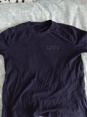 Mörkblå DJUU t-shirt i bomull - Snygg mörkblå t-shirt från DJUU med enkel design och rund hals. T-shirten är kortärmad och tillverkad i mjuk bomull, perfekt för en avslappnad stil. Diskret logga på bröstet ger en clean look. Och priset kan diskuteras ner!