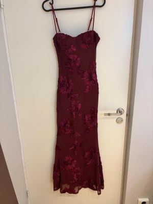Vinröd långklänning med blommor - Vinröd långklänning med tunna axelband och blommigt mönster i samma färgton. Klänningen har figurnära passform och är ärmlös. Den är aldrig använd då jag köpte en annan klänning till balen!