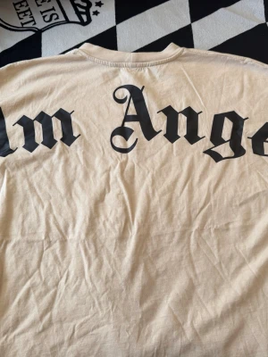Beige Palm Angels t-shirt S - Säljer en beige t-shirt från Palm Angels i storlek S. T-shirten har svart Palm Angels-tryck i gotisk stil både på ryggen och vid kragen. Tillverkad i mjuk bomull och har klassisk passform med korta ärmar.