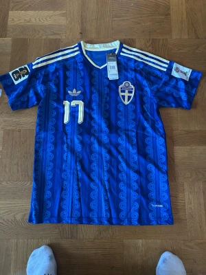 Blå Sverige fotbollströja Adidas - Blå fotbollströja från Adidas med Sveriges landslagsemblem på bröstet och nummer 11 i vitt. Tröjan har vita och guldiga detaljer, klassiska Adidas-ränder på axlarna och mönstrad design. Tillverkad i lätt polyester med AEROREADY-teknologi.