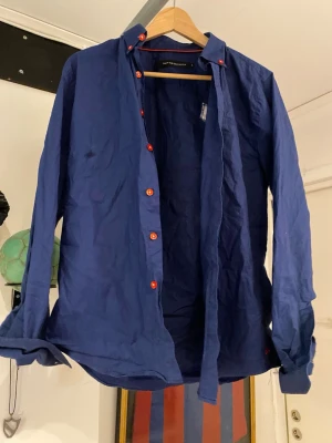 Blå skjorta med röda knappar - Snygg blå skjorta från Scotch & Soda med röda knappar längs hela framsidan. Skjortan har klassisk krage och långa ärmar. Tillverkad i bomull för en skön känsla och avslappnad look.