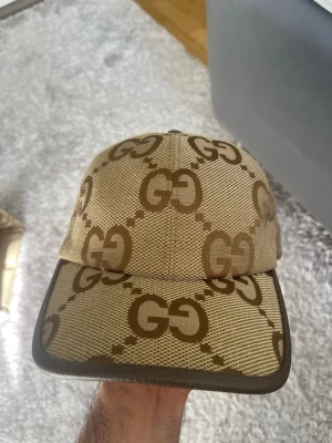 Beige keps från Gucci med GG-mönster - Säljer en beige keps från Gucci med det klassiska GG-monogrammet i brunt över hela kepsen. Kepsen har böjd skärm och justerbart band bak i brunt läder. Tillverkad i slitstarkt tyg som ger en exklusiv känsla och passar perfekt till streetwear-stilen.