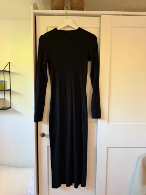 Svart ribbad långklänning - En svart, långärmad långklänning med ribbad struktur. Klänningen har en kroppsnära passform och är tillverkad i ett stretchigt material som ger en snygg siluett. Perfekt för dig som gillar stilrena och minimalistiska plagg.