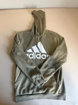Hoodie från Adidas med logga - Snygg grön hoodie från Adidas med stor vit logga på bröstet. Klassisk modell med huva och dragsko samt känguruficka framtill. Perfekt för en avslappnad och sportig stil.
