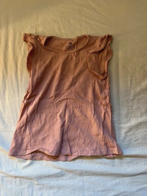 Rosa ärmlös topp från H&M för barn  - Söt ljusrosa topp från H&M i mjuk bomull. Modellen är ärmlös med små volangdetaljer vid axlarna och har en enkel, rak passform. Perfekt för varma dagar och enkel att matcha med olika stilar.