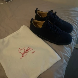 Mörkblåa Louboutin sneakers med röd sula - Stilrena mörkblåa sneakers från Christian Louboutin med klassisk röd sula och svarta skosnören. Ovandelen är i mocka och insidan är beige. Kommer med original dustbag. Perfekta för dig som vill ha en lyxig och tidlös sneaker med ikonisk design.
