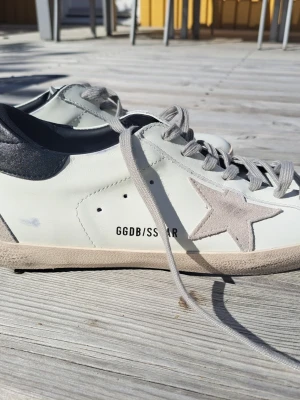 Golden Goose vita sneakers med stjärna - Snygga vita sneakers från Golden Goose med grå mockadetaljer och ikonisk stjärna på sidan. Skorna har grå snörning, svart hälparti och GGDB/SS STAR-tryck på sidan. Tillverkade i läder och mocka för en cool och trendig look.