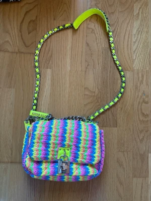 Färgglad axelväska från Steve Madden - Axelväska i regnbågsfärgat tyg med vågigt mönster i neonrosa, gul, grön, blå och lila. Kedjerem med inslag av neonfärgat material och silverdetaljer. Liten metallogo framtill och Steve Madden-märke baktill. Perfekt för dig som gillar färg och statement-accessoarer.