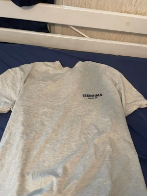 Grå Essentials t-shirt Fear of God - Säljer en grå t-shirt från Essentials Fear of God med svart logotyptryck både fram och bak. T-shirten har klassisk passform och korta ärmar, perfekt för dig som gillar streetwear och enkel stil. Materialet är mjukt och känns skönt mot huden.