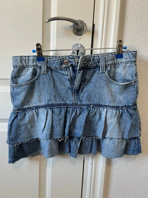 Blå volangkjol i denim Gina tricot  st. 170 - Säljer en blå jeanskjol med volanger och råa kanter. Kjolen har fickor fram, bälteshällor och knappstängning. Perfekt för dig som gillar en avslappnad men trendig stil.