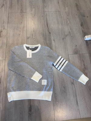 Gråvit stickad tröja Thom Browne - Snygg grå stickad tröja från Thom Browne med vita ribbade muddar, krage och nederkant. Tröjan har fyra vita ränder på ena ärmen och en liten vit patch nertill. Perfekt för dig som gillar stilrena detaljer och klassisk design.