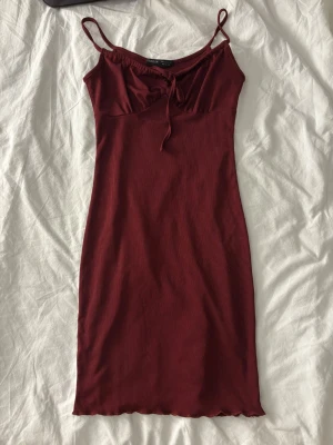 Vinröd ribbad klänning från SHEIN - Vinröd ribbad klänning från SHEIN med smala axelband och knyt framtill. Klänningen är figurnära och har en enkel, stilren design som passar perfekt till varmare dagar. Materialet är mjukt och stretchigt för en bekväm passform.