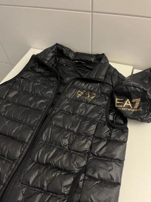 Svart dunväst från Emporio Armani - Snygg svart dunväst från Emporio Armani EA7 med guldfärgad logga på bröstet. Västen har quiltad design, dragkedja framtill och hög krage. Tillverkad i lätt och glansigt material, perfekt för lager-på-lager och streetwear-stil.