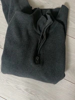 Mörkgrå ribbad ziptröja Dobber - Mörkgrå ribbstickad tröja från Dobber med halv dragkedja framtill. Tröjan har hög krage och är tillverkad i ett mjukt, stretchigt material som ger en skön passform. Perfekt för lager på lager och enkel att matcha med olika outfits.