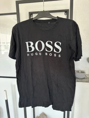 Svart Hugo Boss t-shirt med logga - Snygg svart t-shirt från Hugo Boss med stor vit logga på bröstet. Klassisk rund halsringning och korta ärmar. Tillverkad i mjuk bomull som är skön att bära. Perfekt för en stilren och avslappnad look.