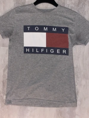 Grå Tommy Hilfiger t-shirt - Snygg grå t-shirt från Tommy Hilfiger med stort logotryck framtill i vitt, marinblått och rött. Klassisk rund halsringning och korta ärmar. Tillverkad i mjuk bomull för skön känsla och avslappnad passform.