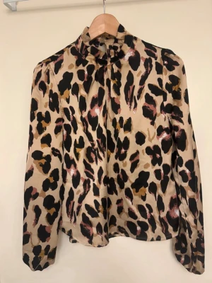 Leopardmönstrad blus med hög krage - Snygg blus i beige med leopardmönster i svart, brunt och rosa toner. Hög krage med knapp i nacken. Tillverkad i ett glansigt, tunt material som ger en lyxig känsla. Perfekt för dig som vill sticka ut med ett trendigt mönster.