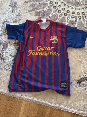 FC Barcelona Messi matchtröja - Säljer en klassisk FC Barcelona matchtröja från Nike med röda och blå vertikala ränder, Qatar Foundation-tryck på bröstet och Messi 10 på ryggen. Tröjan har klubbmärke och Nike-logga framtill samt korta ärmar. Perfekt för fotbollsfans!