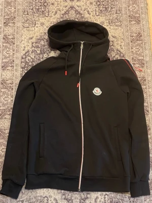 Svart hoodie från Moncler med dragkedja - Svart hoodie från Moncler med hel dragkedja, röda detaljer på snören och axlar samt Moncler-logga på bröstet. Mjuk bomullskänsla, två fickor fram och klassisk huva. Perfekt för en stilren och sportig look.