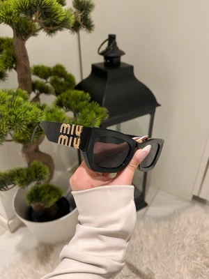 Svarta Miu Miu solglasögon med guldlogo - Snygga svarta solglasögon från Miu Miu med breda bågar och stora, mörka glas. Sidorna pryds av en stor guldig Miu Miu-logo som ger en lyxig känsla. Perfekt accessoar för att lyfta din stil.