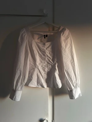 Vit blus med puffärm från Vero Moda - Stilren vit blus från H&M med puffiga långärmar och bred, rundad urringning. Blusen har knappar framtill och breda manschetter vid ärmsluten. Tillverkad i ett lätt bomullstyg som ger en luftig känsla.