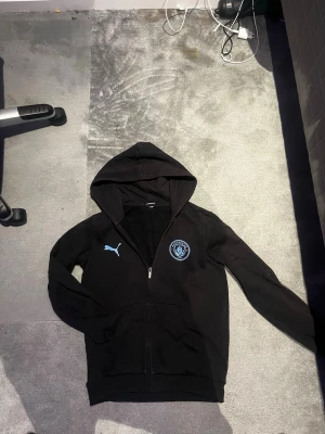Svart Manchester City hoodie Puma - Svart hoodie med dragkedja från Puma och Manchester City-logga på bröstet. Klassisk huva och två fickor framtill. Perfekt för dig som gillar sportig stil och vill visa ditt lagstöd. Materialet är mjukt och bekvämt, med diskret blå Puma-logga.
