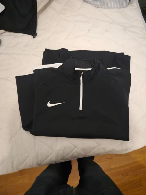 Svart Nike Dri-Fit tröja - Säljer ett svart tröja från Nike med Dri-Fit-teknologi. Setet består av en långärmad tröja med halv dragkedja och vita detaljer samt matchande byxor. Perfekt för träning eller sportaktiviteter. Snygg och sportig design med klassisk Nike-logga.