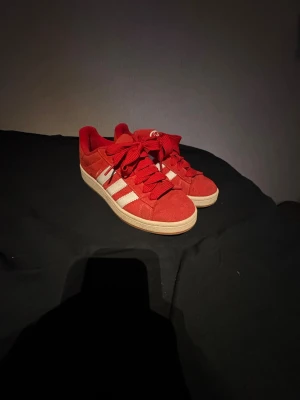 Röda Adidas sneakers i mocka - Snygga röda Adidas sneakers i mocka med klassiska vita ränder på sidorna och vit sula. Skorna har röda skosnören och låg profil som ger en clean och sportig look. Perfekta för dig som vill sticka ut med färg och stil. Knappt använt dem kan även tvätta de om någon är bra intresserad 