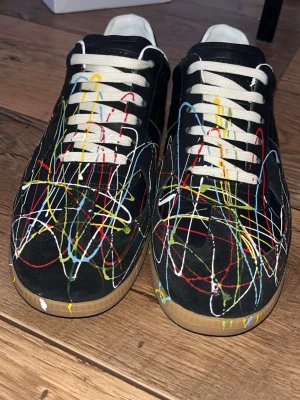 Svarta sneakers med färgstänk - Unika svarta MAISON margiella gats med coolt färgstänksmönster i rött, blått, gult och vitt. Skorna har vita snören, vit häl och klassisk gummisula. Perfekt för dig som vill sticka ut med en kreativ och konstnärlig vibe. Materialet är en mix av läder och textil.