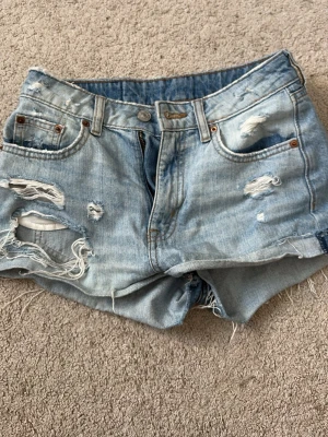 H&M Ljusblå jeansshorts - Säljer ett par ljusblå jeansshorts med slitna detaljer och fransiga kanter. Shortsen har klassisk femficksdesign, knapp och dragkedja framtill. Perfekta för en avslappnad och trendig look.