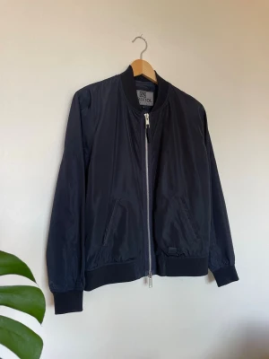 Bristol Bomberjacket Marinblå L - Bäst färgåtergivning på första bilden och sista bilden!  Säljer en klassisk och stilren bomberjacka från det svenska märket Brixtol Textiles. Modellen heter Harris och är en av deras mest uppskattade basplagg tack vare sin tidlösa design och höga kvalitet.  Jackan är i en mycket snygg mörkblå (navy) nyans som passar till allt. Den har de klassiska bomber-detaljerna: ribbstickad krage, muddar vid ärmslut och nederkant, samt rejäla dragkedjor.  Färgen är fortfarande djup och mörk, helt utan tecken