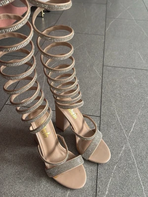 Beige gladiatorsandal med strass - Säljer ett par beige gladiatorsandaler från Simmi med höga blockklackar och flera remmar dekorerade med strass. Sandalerna har öppen tå och en trendig design som snörs upp längs benet. Perfekta för dig som vill sticka ut med din stil.