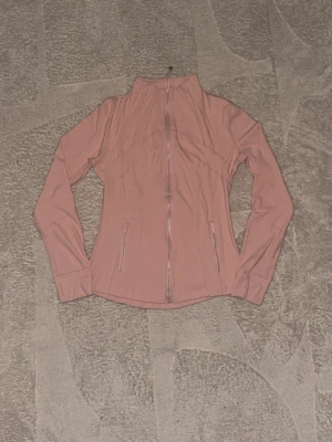 Lululemon Define Jacket - Säljer en ljusrosa lululemon define jacket med hel dragkedja framtill och två fickor med dragkedja. Jackan har lång ärm, hög krage och figurnära passform. Perfekt för träning eller utomhusaktiviteter. Materialet känns stretchigt och bekvämt.